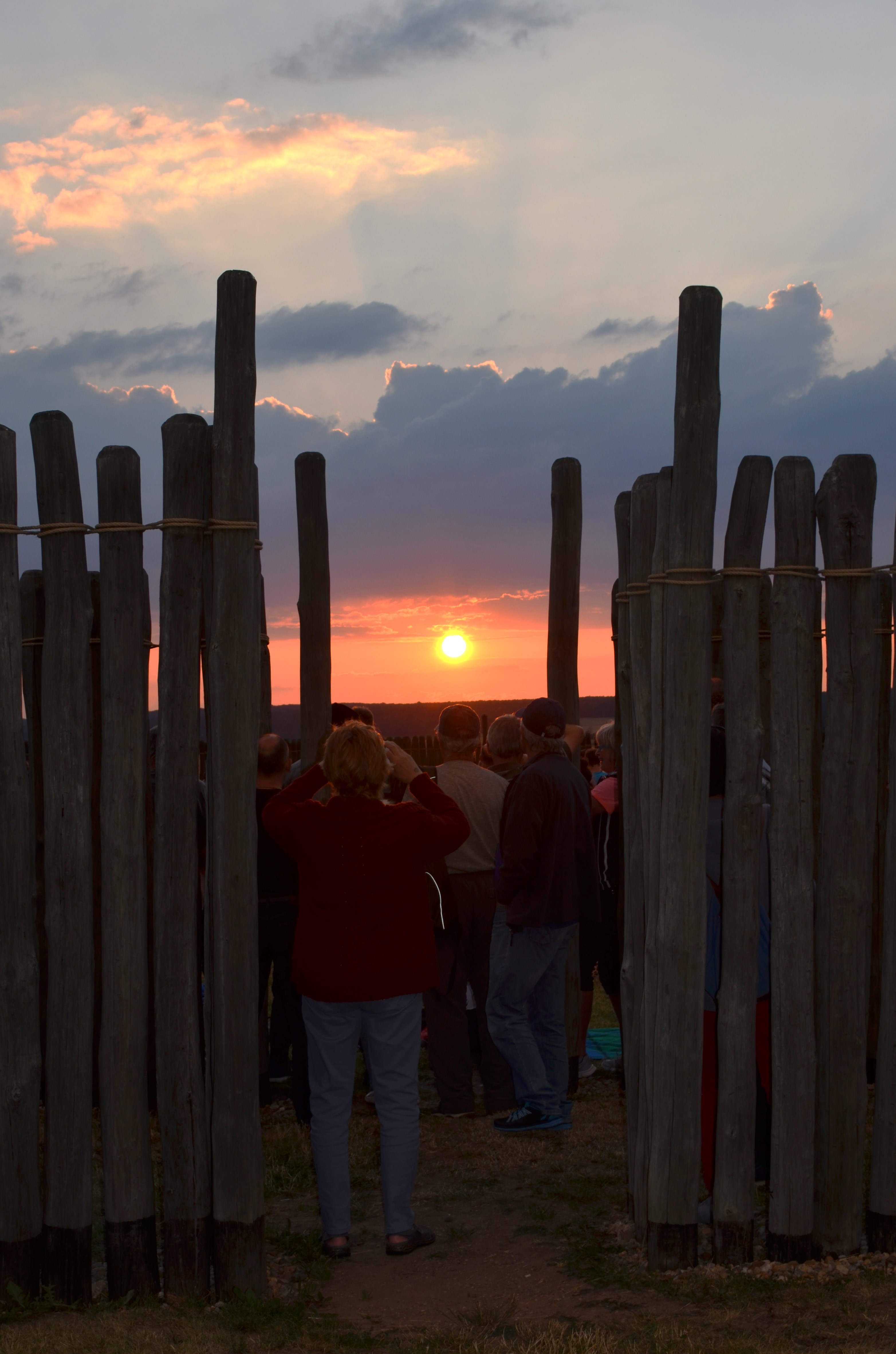 Blick durch eine Lücke zwischen als Palisaden aufgerichtete Holzpfosten hindurch zum Sonnenuntergang. Einige Personen stehen mit dem Rücken zum Betrachter und beobachten den Sonnenuntergang durch die Lücke. 